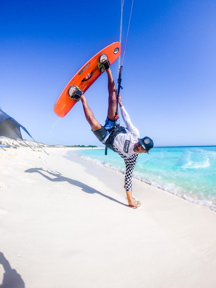Energetic kiteboarder performing a stunt on a sunny beach with clear blue skies and ocean in the background.